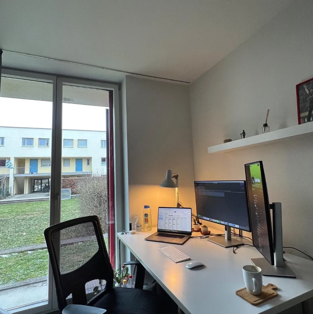 A tidy desk by a window: laptop, a horizontal and a vertical monitor, a lamp and a coffee mug